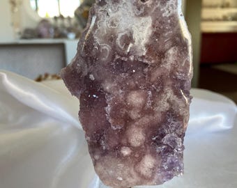 May include: A large, raw amethyst crystal geode, displaying shades of purple and white, mounted on a black metal stand. The crystal has a rough, textured surface with visible crystalline structures. The geode is displayed on a white satin cloth.