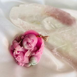 Peut inclure: Un pendentif rose sculpté en forme de Bouddha rieur. Le pendentif est doté d'une bélière dorée ornée de petites pierres transparentes. Le Bouddha est détaillé avec des traits sculptés et des accents verts et blancs. Le pendentif est présenté sur un tissu blanc et soyeux.