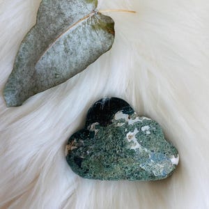 May include: A green and white agate stone shaped like a cloud, resting on a white fluffy surface.