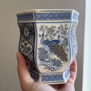 Op de afbeelding: Een blauw-witte keramische vaas met een gouden rand en accenten. De vaas heeft een pauwenontwerp met bloemen- en geometrische patronen. De vaas wordt in een hand gehouden.