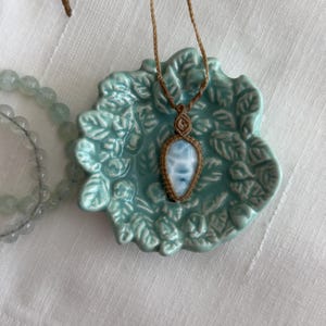 Puede incluir: Un collar de macramé artesanal con un colgante de piedra azul claro y blanco. El colgante está engarzado en un cordón marrón tejido y cuelga de un cordón a juego. El collar descansa sobre un plato de cerámica azul claro con un diseño de hojas.