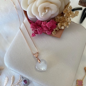 Könnte beinhalten: Eine zarte Halskette aus Roségold mit einem weißen herzförmigen Anhänger. Die Halskette wird in einer weißen Schachtel mit einem rosa-weißen Blumenarrangement präsentiert.