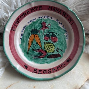May include: A round ceramic plate with a scalloped green edge and a pink border. The center features a green painted area with illustrations of carrots, a shrimp, an artichoke, and tomatoes. Text reads RISTORANTE MANARINI ANTIPASTI MAGRI ASSORTITI ALLA MANARINI BERGAMO.