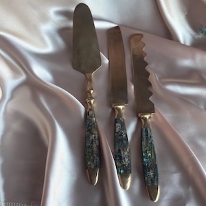 Peut inclure: Un ensemble de trois ustensiles de service : une pelle à gâteau, un couteau à beurre et un couteau dentelé. Chaque ustensile a une lame en laiton et un manche incrusté de nacre irisée. L'ensemble est présenté sur une surface satinée.