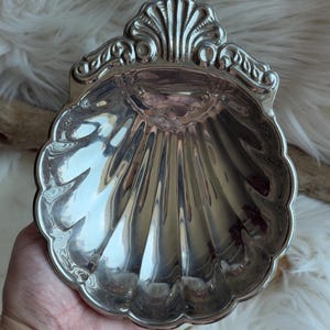 Pode incluir: Um prato de metal prateado polido em forma de concha de vieira. O prato apresenta um design decorativo em relevo em forma de leque na borda superior e sulcos verticais profundos que irradiam do centro para a borda festonada.
