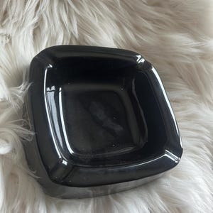 Puede incluir: Un cenicero cuadrado de color negro brillante con esquinas redondeadas y cuatro muescas distintivas a lo largo del borde. El cenicero presenta una superficie lisa y reflectante y un centro profundo y abierto, hecho de un material s&oacute;lido.