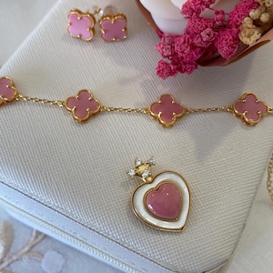 May include: A jewelry set featuring a gold necklace and earrings with pink clover-shaped charms. A heart-shaped pendant with a crown is also included. The jewelry is displayed on a white surface with pink flowers in the background.