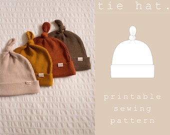 Puede incluir: Un patrón de costura imprimible para un gorro de lazo para bebé. El patrón se muestra con un contorno blanco sobre un fondo marrón claro. El texto "baby tie hat." está por encima del patrón y el texto "printable sewing pattern" está por debajo del patrón. El texto "patterns by HUM apparel" está en la parte inferior de la imagen.