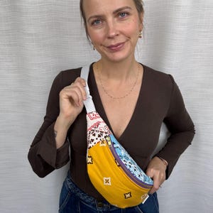 May include: A crossbody fanny pack featuring a patchwork design of yellow, light blue, and white fabric with black bandana-style patterns. The bag includes a white adjustable strap, a gold-toned zipper, and a small rectangular brand label on the side.