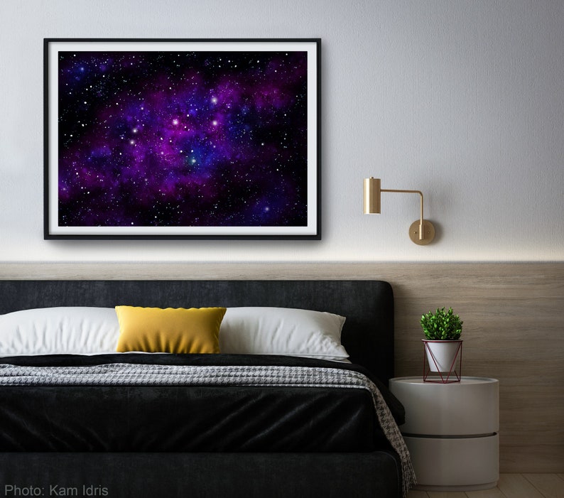 May include: A framed print of a galaxy with purple and blue nebulae and white stars against a black background. The print is hanging on a white wall above a black headboard with a yellow pillow and white bedding.