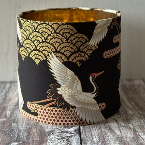 May include: Cylindrical lampshade with a black background and a gold interior. The exterior features a pattern of white cranes, gold wave-like designs.