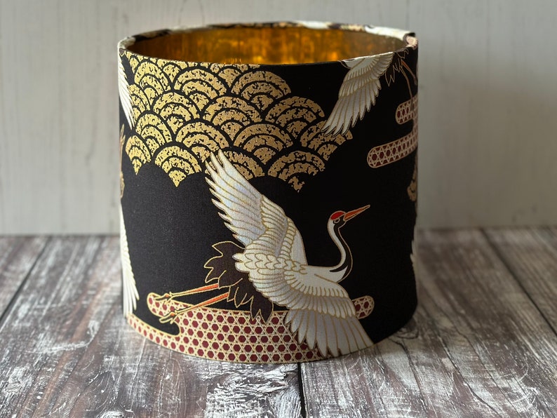 May include: Cylindrical lampshade with a black background and a gold interior. The exterior features a pattern of white cranes, gold wave-like designs.