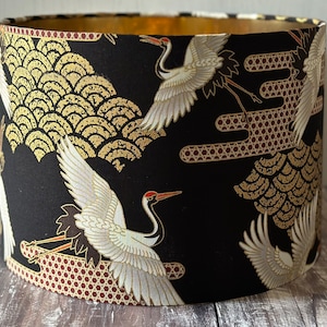 May include: Drum lampshade with a black base, decorated with a gold and white crane pattern. The cranes have red details, set against gold wave motifs and geometric shapes. The inside of the shade is gold.