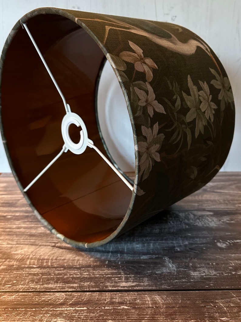 May include: A green lampshade with a floral pattern and a white bird design. The lampshade has a brown lining and a white metal frame.