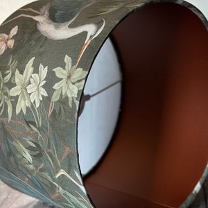May include: A green lampshade with a floral pattern featuring a heron and white flowers. The inside of the lampshade is a solid brown color.