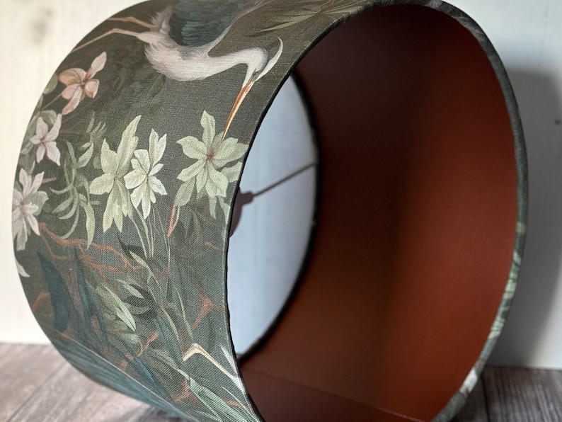May include: A green lampshade with a floral pattern featuring a heron and white flowers. The inside of the lampshade is a solid brown color.