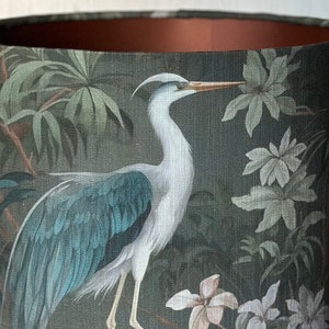 May include: A green lampshade with a white heron and floral pattern. The heron has a long neck and blue wings. The flowers are white with green leaves.