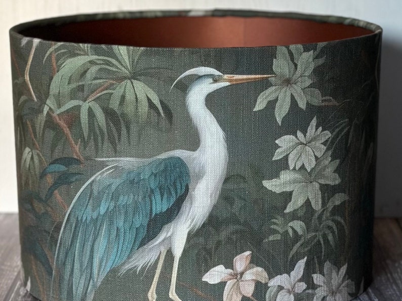 May include: A green lampshade with a white heron and floral pattern. The heron has a long neck and blue wings. The flowers are white with green leaves.