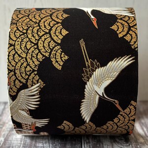 May include: A fabric-covered item with a black backdrop, adorned with gold and white crane illustrations. The design incorporates gold, wave like patterns. The cranes have white wings, red accents, and are depicted in flight. The item is cylindrical.