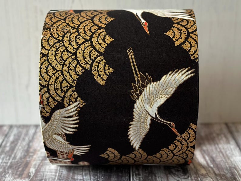 May include: A fabric-covered item with a black backdrop, adorned with gold and white crane illustrations. The design incorporates gold, wave like patterns. The cranes have white wings, red accents, and are depicted in flight. The item is cylindrical.