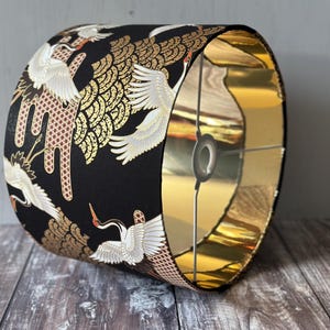 May include: A cylindrical lampshade with a black exterior featuring a pattern of white cranes with gold accents. The interior is gold. The lampshade is designed to fit over a light fitting.