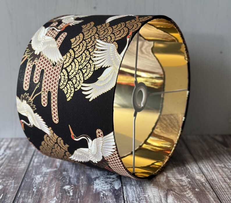 May include: A cylindrical lampshade with a black exterior featuring a pattern of white cranes with gold accents. The interior is gold. The lampshade is designed to fit over a light fitting.