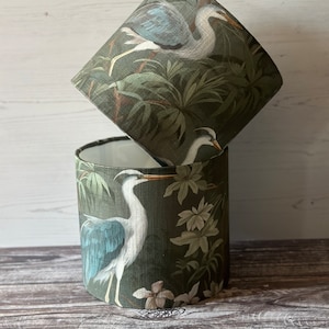 May include: Two green lampshades with a white heron and tropical foliage design. The lampshades are stacked on top of each other.