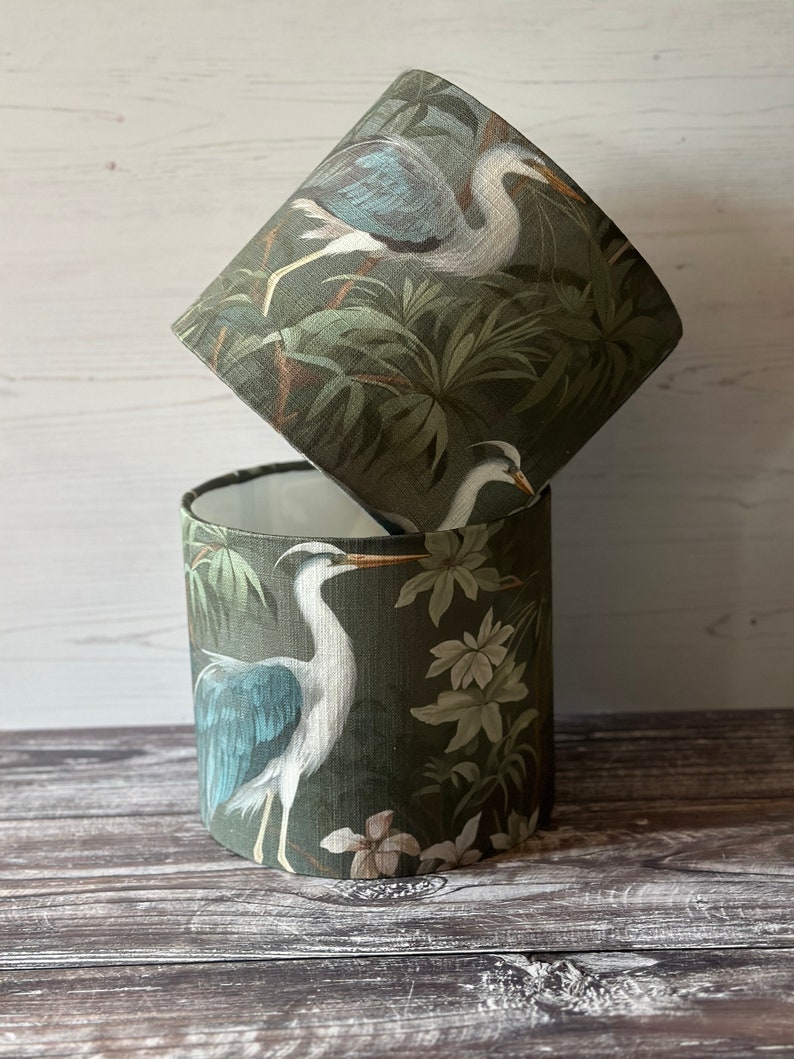 May include: Two green lampshades with a white heron and tropical foliage design. The lampshades are stacked on top of each other.