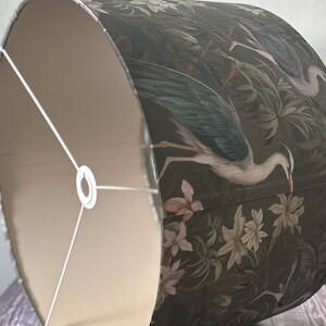 May include: A drum-shaped lampshade with a white interior and a dark green exterior. The exterior features a floral pattern with white birds and leaves.