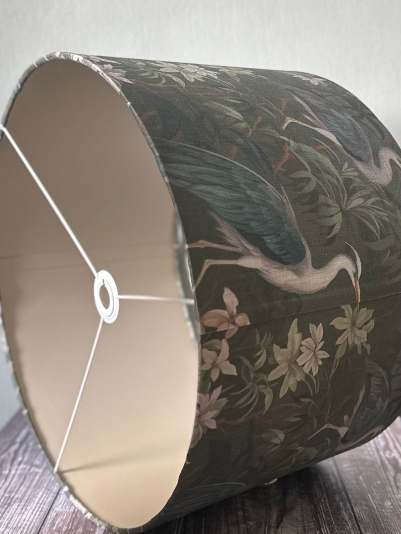 May include: A drum-shaped lampshade with a white interior and a dark green exterior. The exterior features a floral pattern with white birds and leaves.