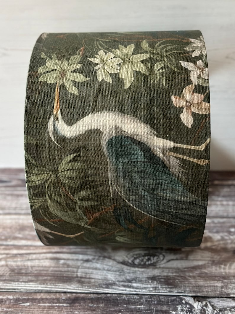 May include: A green lampshade with a white heron and floral pattern. The heron is standing in a swamp with green foliage and white flowers.