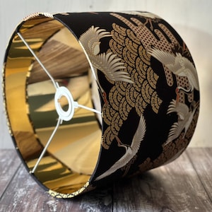 May include: A cylindrical lampshade with a black exterior featuring a gold and white crane design. The interior is gold, and the frame is white.