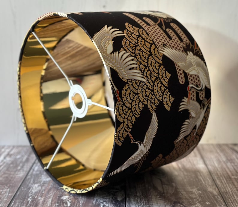 May include: A cylindrical lampshade with a black exterior featuring a gold and white crane design. The interior is gold, and the frame is white.