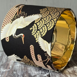 May include: A cylindrical lampshade with a black background and a gold interior. The exterior features a repeating pattern of white cranes with red beaks and gold accents.