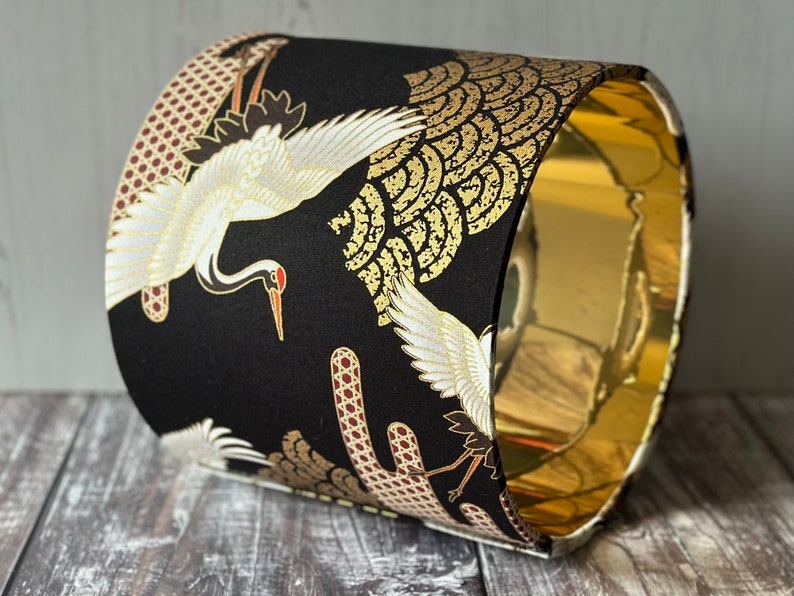 May include: A cylindrical lampshade with a black background and a gold interior. The exterior features a repeating pattern of white cranes with red beaks and gold accents.