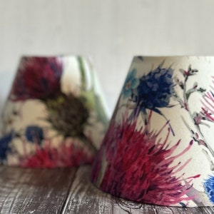 May include: Two white lampshades with a floral pattern featuring pink, blue, and green watercolour flowers. The lampshades are on a wooden surface.