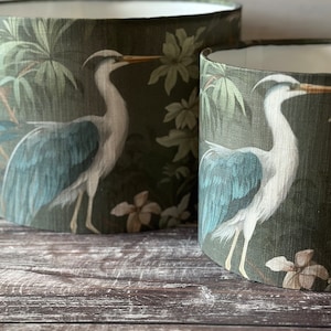 May include: Two green lampshades with a white heron design and tropical foliage. The lampshades are on a wooden surface.