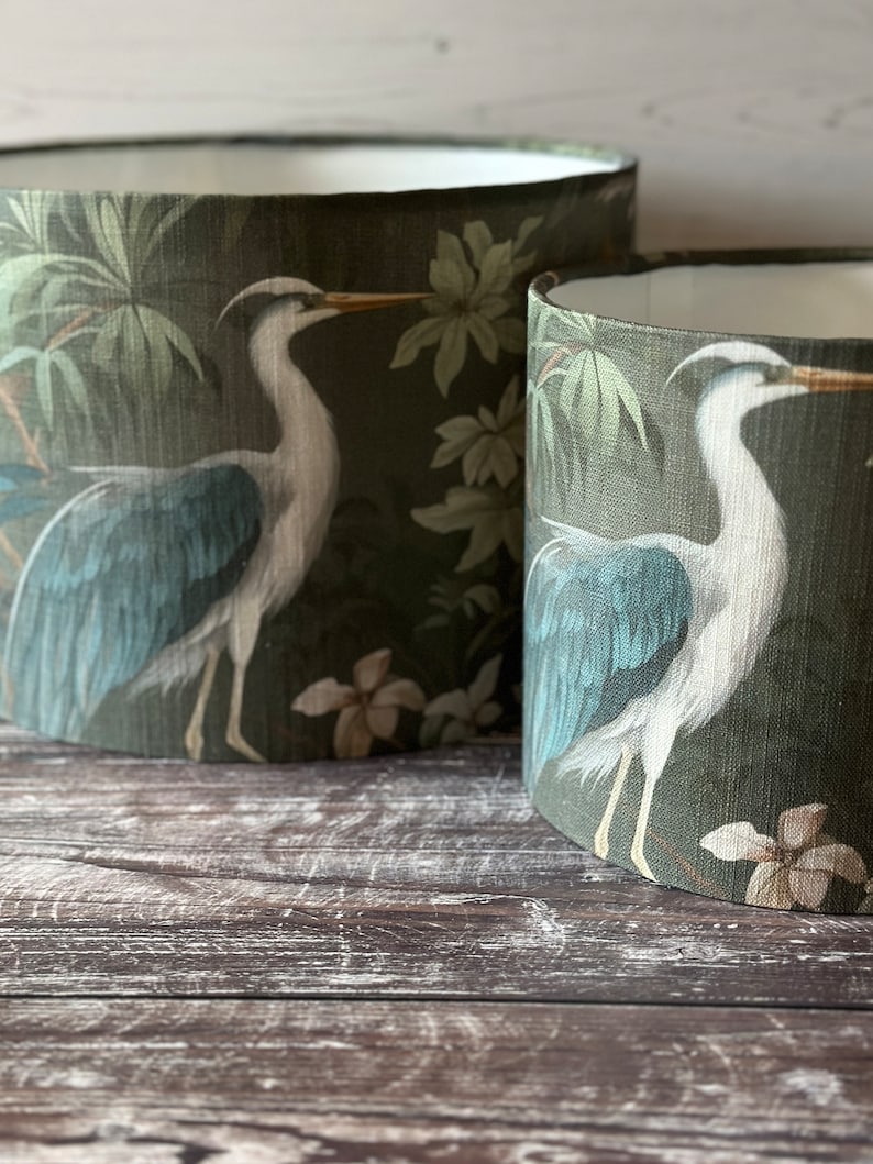 May include: Two green lampshades with a white heron design and tropical foliage. The lampshades are on a wooden surface.