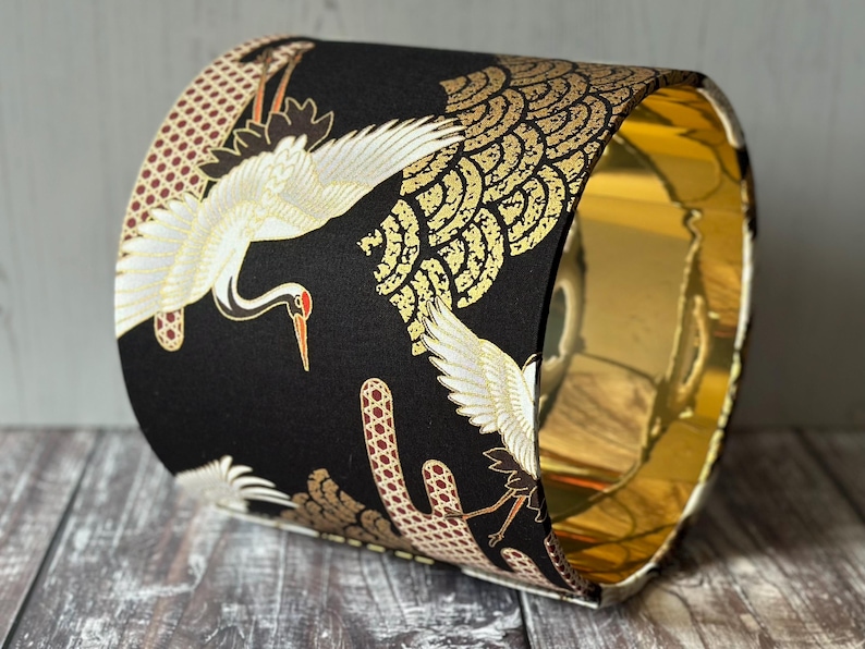 May include: A cylindrical lampshade with a black exterior featuring white cranes with red beaks and gold accents. The interior is gold. The lampshade is a decorative lighting element.