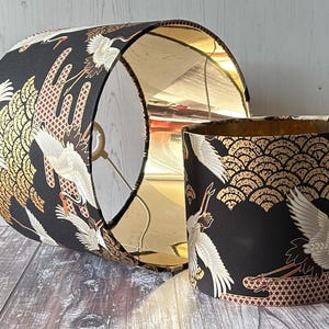 May include: Two black lampshades with a gold interior and a Japanese crane design. The exterior features white cranes, gold fan-shaped patterns.