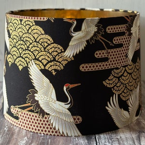 May include: Cylindrical lampshade featuring a black background with a gold and white crane design. The interior is gold. The pattern includes gold wave motifs. A decorative lighting element.