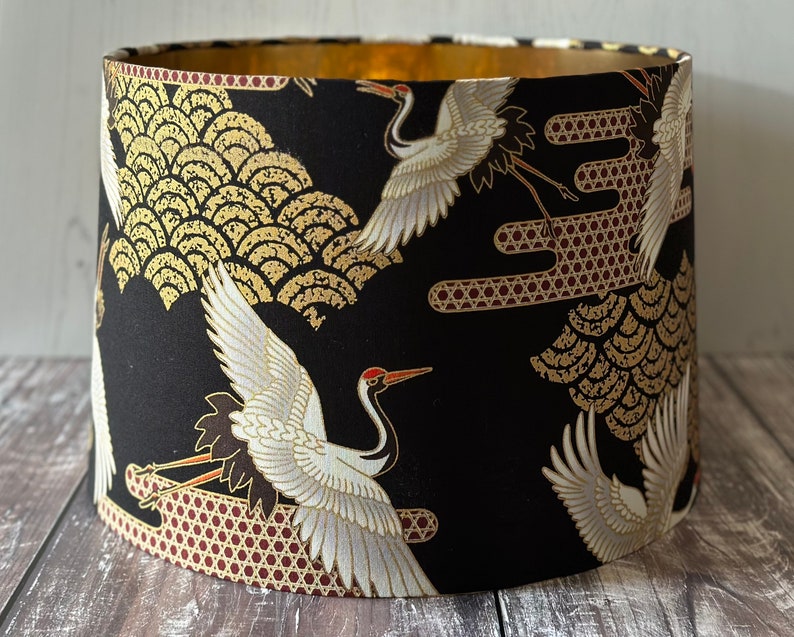 May include: Cylindrical lampshade featuring a black background with a gold and white crane design. The interior is gold. The pattern includes gold wave motifs. A decorative lighting element.