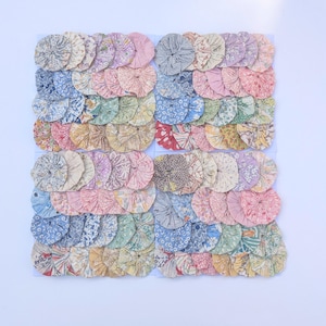 May include: A collection of numerous circular fabric yo-yos arranged in a grid. Each yo-yo features a unique floral or geometric pattern in various pastel and vibrant colours, including shades of blue, pink, yellow, green, and grey, with gathered centres.