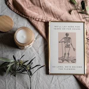May include: A gold framed print with a black and white illustration of a skeleton holding a stick. The print has a pink background and the text "We'll lay here for years or for hours. So long, we'd become the flowers."