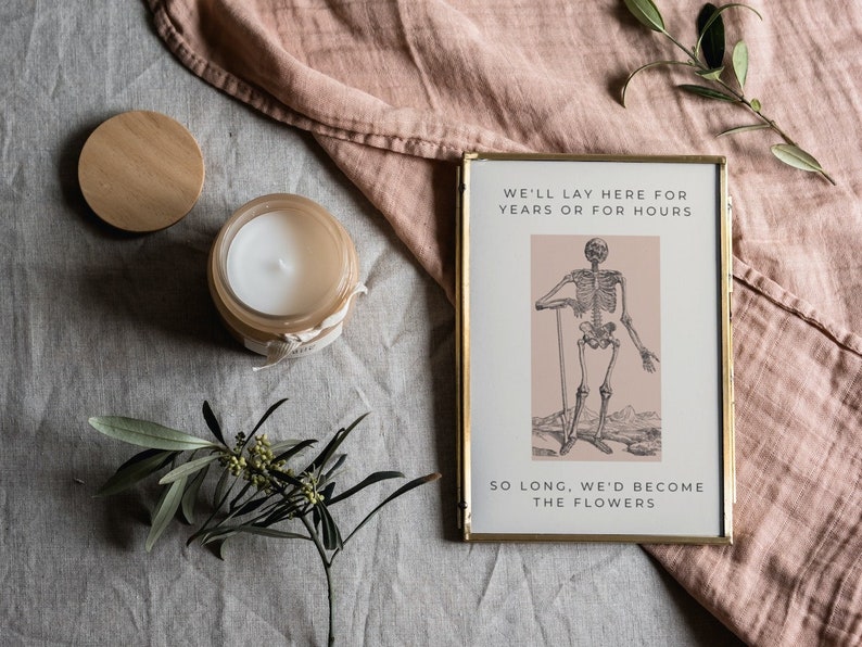 May include: A gold framed print with a black and white illustration of a skeleton holding a stick. The print has a pink background and the text "We'll lay here for years or for hours. So long, we'd become the flowers."