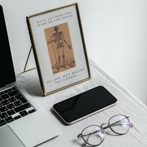 May include: A gold-framed picture with a black and white illustration of a skeleton standing with a stick. The text in the picture reads "We'll lay here for years or for hours. So long, we'd become the flowers."