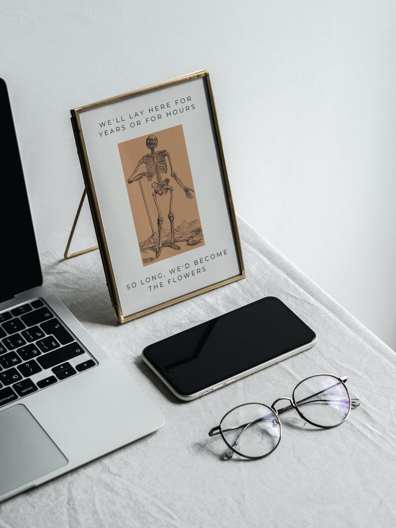 May include: A gold-framed picture with a black and white illustration of a skeleton standing with a stick. The text in the picture reads "We'll lay here for years or for hours. So long, we'd become the flowers."