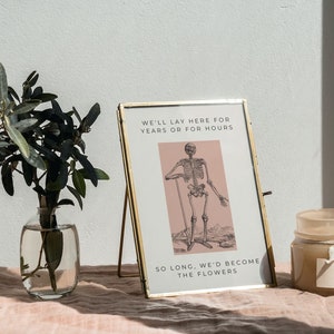 May include: A gold-framed picture with a black and white illustration of a skeleton standing on a pink background. The text in the picture reads "We'll lay here for years or for hours. So long, we'd become the flowers."