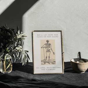 May include: A framed print featuring a black and white illustration of a skeleton holding a stick. The text on the print reads "We'll lay here for years or for hours. So long, we'd become the flowers."