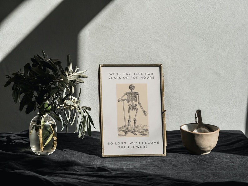 May include: A framed print featuring a black and white illustration of a skeleton holding a stick. The text on the print reads "We'll lay here for years or for hours. So long, we'd become the flowers."